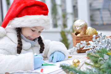 Little happy girl in a white fur coat, gloves and a santa hat is writing a letter to Santa Claus on the background of a Christmas house outdoors. Happy new year and Merry christmas!