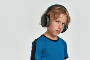 Portrait of tired little sportive boy child in sportswear and headphones looking at camera while posing, standing isolated over white background