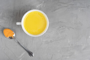 Obraz na płótnie Canvas Golden turmeric milk or haldi doodh which is source of antioxidants and anti-inflammatory compounds served in cup with spoon of curcuma powder on gray concrete background. Image with copy space