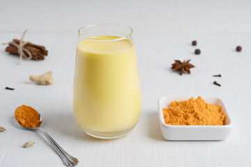 Ayurvedic Indian golden turmeric milk or haldi doodh which is source of antioxidants and anti-inflammatory compounds in drinking glass with curcuma powder on white wooden background. Horizontal