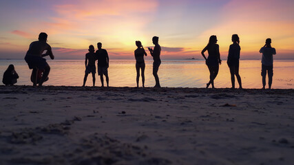 Silhouettes of people on the sandy beach enjoying the sunset and taking pictures on smartphones against the background of the sea