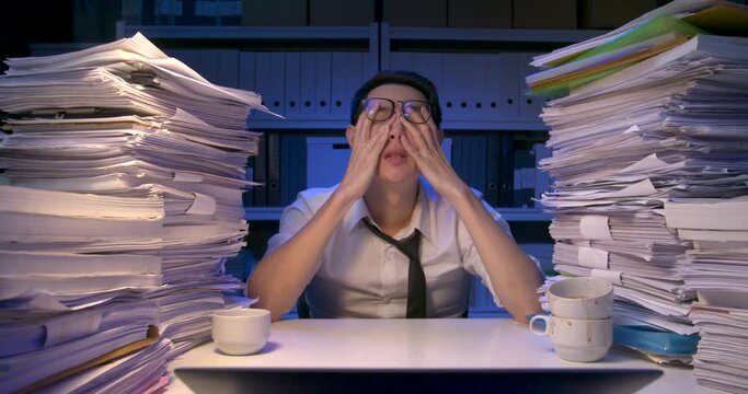Asian business man is sitting at desk covered with stack of paperwork and feeling fatigued after working hard at late night. Tired alone male break for rest, take off glasses and massage eye, temples.