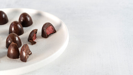 Homemade dark chocolate sweets on white plate. Raspberry filling in candy. One candy cut in half. Selective focus. Side view.