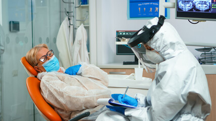 Dentist assistant in coverall interrogating senior woman during global coronavirus pandemic writing...