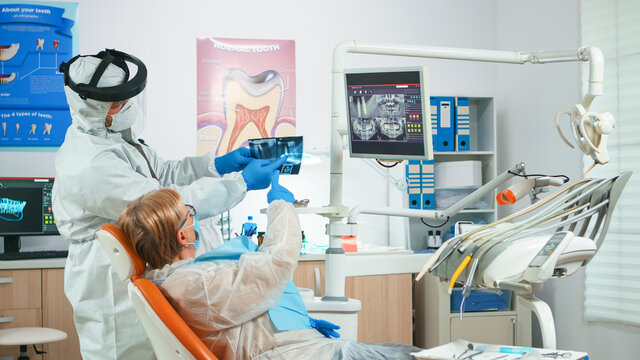 Dentist with face shield reviewing panoramic mouth x-ray image to patient during global pandemic. Assistant and doctor talking with senior woman wearing suit, coverall, protection suit, mask, gloves