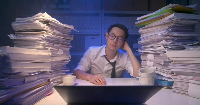 Exhausted asian business man is holding head with hand and falling asleep. Sleepy alone male dozing off on desk covered with stack of paperwork while working hard at late night. Smoke on background.