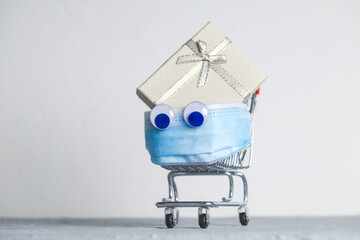 Funny creative shopping cart with protective mask and Christmas gift box. Safety Christmas shopping during corona virus pandemic.