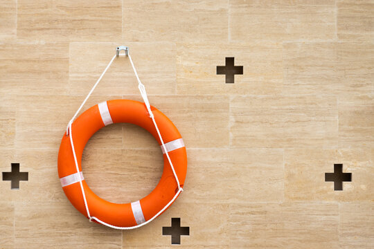 Lifebuoy Ring Orange Color With White Rope Life Saving Equipment To Be Throw To A Person In Water, Sea And Pool Hanging On Light Brown Granite Wall And Cross Holes Pattern Background