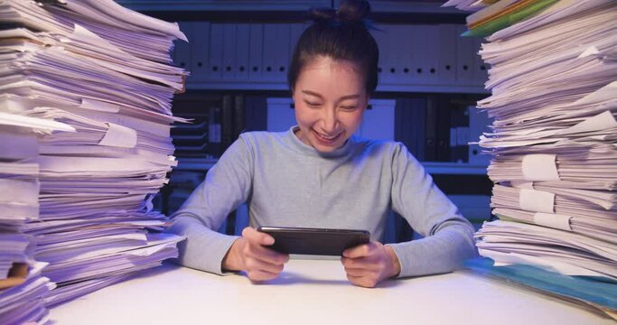 Relaxing asian business woman is sitting at desk covered with stack of paperwork at home and playing mobile game while working hard overload at late night. Exciting alone girl enjoy happy and fun.
