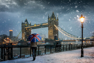 Londoner Winterkonzept mit einem Touristen, der einen Souvenir-Regenschirm mit britischer Flagge hält, der während der Abendzeit mit Schneefall und kalten Temperaturen vor der Tower Bridge steht, Vereinigtes Königreich
