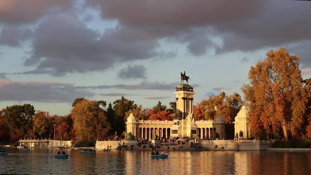 Pond with boats in Retiro Park of Madrid and a monument with columns in the background during sunset in a cloudy day of autumn