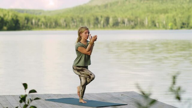 Young Caucasian woman in sportswear doing eagle pose on mat while practicing yoga on pier near scenic lake on summer day