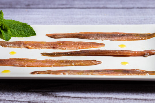 Tapa de anchoa gourmet con puntitos de aceite en un plato blanco rectangular y una tabla r&uacute;stica gris claro de fondo