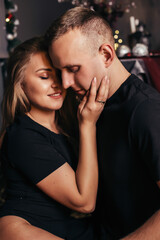 View of a passionate romantic couple spending time before New Year at home with garlands. Young woman and handsome man hug, kiss. Christmas time.