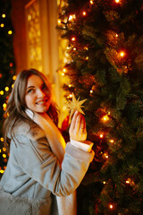 Beautiful woman posing  by the christmas tree at winter holidays. Young woman wearing stylish winter coat,  scarf. Christmas and New Year magic. Lights around.