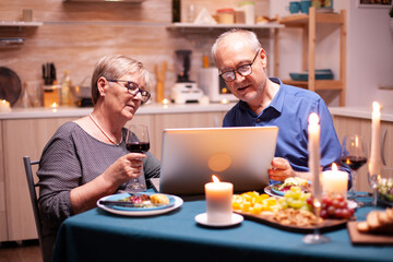 Elderly couple using laptop in the kitchen celebrating relationship. Senior people sitting at the table browsing, searching, using laptop, technology, internet, celebrating their anniversary in the