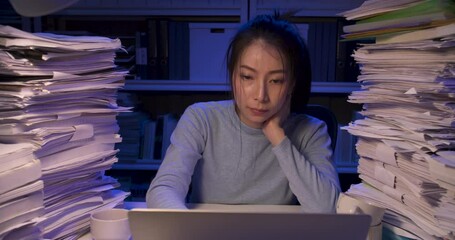 Serious asian business woman is sitting at desk covered with stack of paperwork and typing on keyboard notebook. Unhappy alone girl tired and sighting in frustration while working hard at late night.