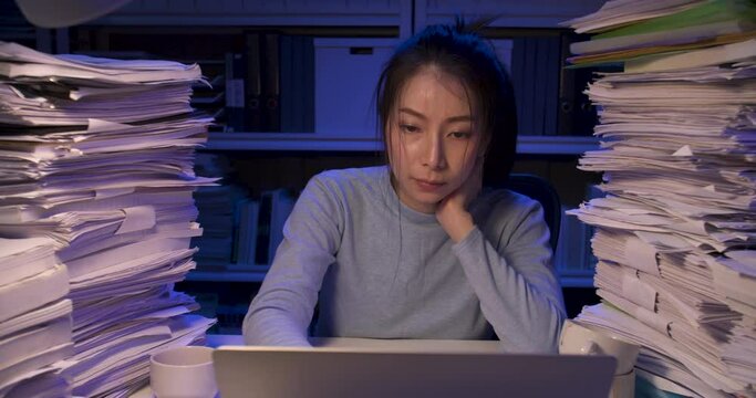 Serious asian business woman is sitting at desk covered with stack of paperwork and typing on keyboard notebook. Unhappy alone girl tired and sighting in frustration while working hard at late night.