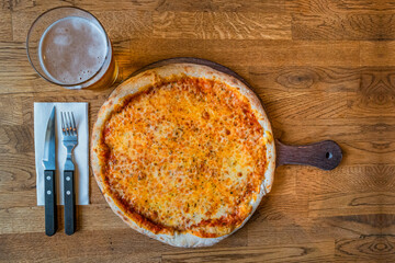 Delicious and appetizing fresh baked pizza margarita with cheese and tomato served with glass of beer and fork and knife with wooden background, details, closeup.