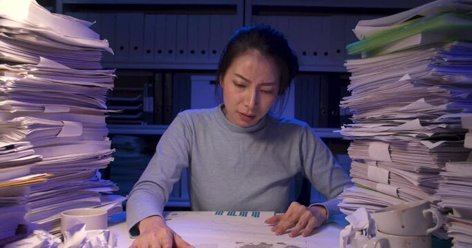 Stressed asian business woman is sitting at desk covered with stack of paperwork and thinking then being struck. Angry alone girl crumpling paper and throws it away while working hard at late night.