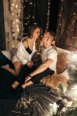 Passionate couple sitting on the bed near the Christmas tree. Real emotions. Joyful moments of comfort during the winter holidays. Christmas romantic mood