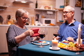 Senior woman looking surprised at gift box from husband during dinner in kitchen. Happy cheerful elderly couple dining together at home, enjoying the meal, celebrating their marriage , surprise