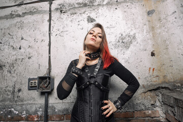 A  attractive dominant woman with piercings and bright hair in a black corset, with leather harnesses and bracelets posing against the background of an old   wall. - obrazy, fototapety, plakaty