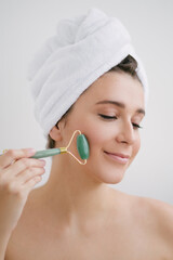 Beautiful young woman with perfect skin wearing towel on head using a jade face roller with natural quartz stones.