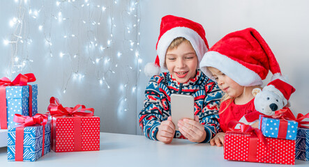 Online christmas greetings. Brother and sister in Santa hats smile and talking on the phone.
