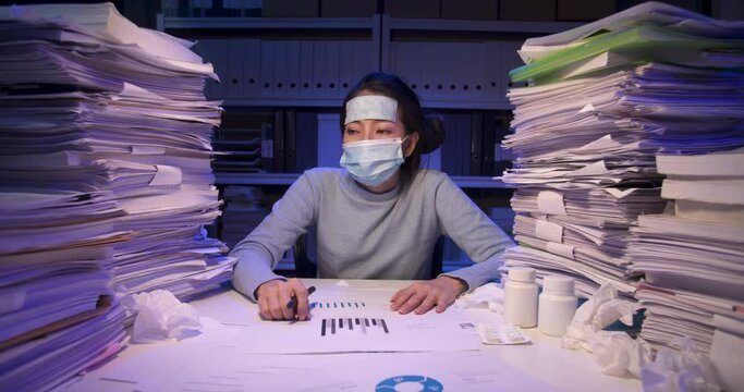 "Sick asian business woman is sitting at desk covered with stack of paperwork and have cooling gel sheet on head, wears face mask. Alone girl coughing and running nose while working hard at late night