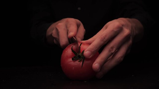 Men's hands cut a red tomato into 2 parts on a dark background