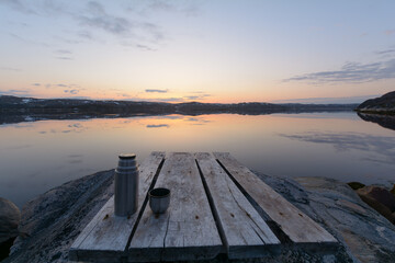 Fototapeta na wymiar Sunset. Calm water in the bay. Wooden pier. Tea in a thermos is on the pier.