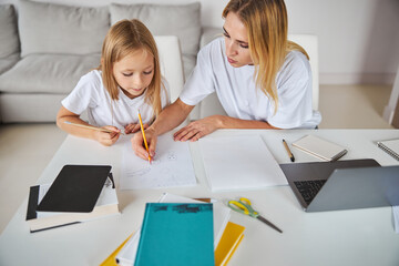 Caring mother in casual clothes working with daughter while sitting in home office