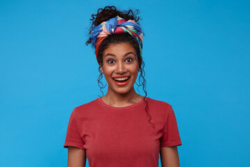 Agitated young attractive brunette curly woman with casual hairstyle raising surprisedly eyebrows while looking excitedly at camera, isolated over blue background