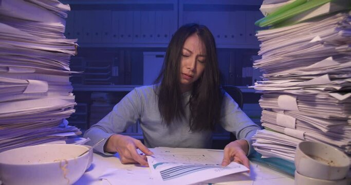 Sleepy exhausted asian business woman looking at document and mess of her hair while working hard at late night. Stressed alone girl throw papers and lay down on desk covered with stack of paperwork.