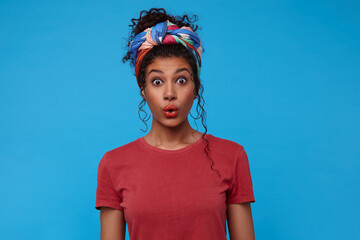 Bemused young attractive dark haired curly woman with multi-colored headband rounding surprisedly her brown eyes while posing against blue background