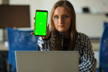 Girl with glasses holding a phone with a green screen sitting at a laptop.