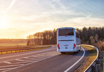 A passenger bus travels along the highway against the backdrop of a forest and a sunset in the sky....