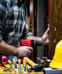 Close-up. Carpenter with an electric screwdriver fixes the lock of a wooden door. Construction...