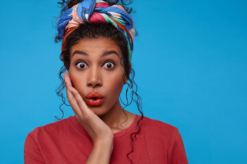 Indoor shot of young bemused dark haired curly woman keeping raised hand on her face while looking at camera with wide eyes opened, isolated over blue background