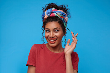 Portrait of young pretty brunette curly female looking cheerfully aside with broad smile and playing with her hair, standing over blue background with raised hand