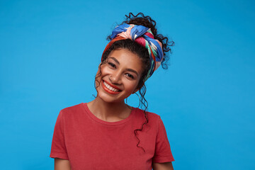 Happy young cute brunette curly female with nose piercing showing her pleasant emotions and smiling cheerfully while looking at camera, isolated over blue background