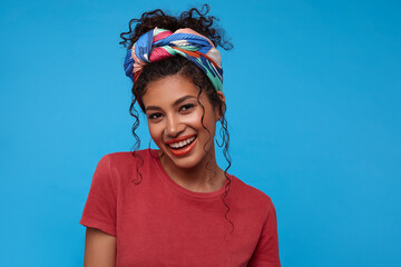Beautiful young pretty dark haired curly woman with casual hairstyle looking gladly at camera and smiling broadly while standing over blue background