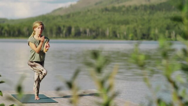 Young blonde woman in sportswear practicing eagle pose on mat while having outdoor yoga workout on pier near beautiful lake on summer day