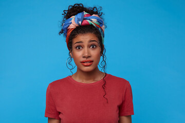 Bewildered young attractive dark haired curly woman with multi-colored headband frowning eyebrows while looking confusedly at camera, isolated over blue background