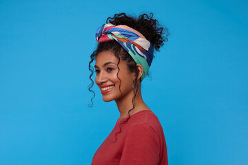 Side view of young cheerful brunette curly woman with festive makeup smiling broadly while looking gladly ahead, isolated over blue background in casual clothes