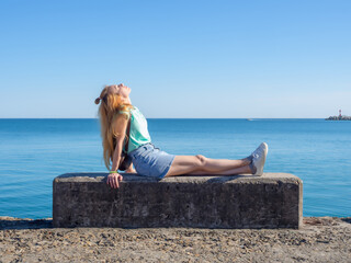 A blonde girl sits on the background of the sea on a concrete slab stretching her legs