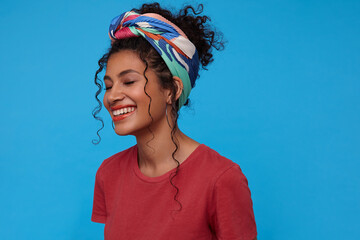 Good looking happy young brunette curly woman with multi-colored headband being in nice mood and miling gladly with closed eyes while standing over blue background