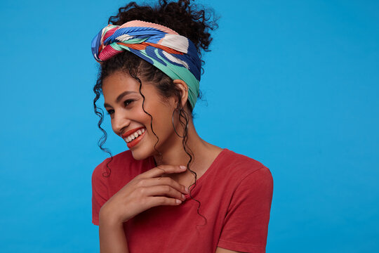 Happy young lovely brown haired curly female with gathered hair smiling happily and touching her neck with raised hand, isolated over blue background