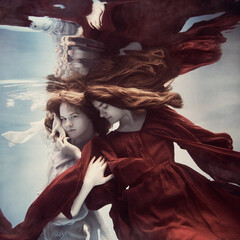 Two girls underwater in blue and red dresses with developing long hair seem to fly in...